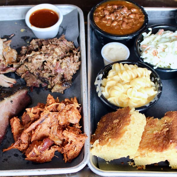 Two metal trays filled with Fat Pete's BBQ meats, sides like mac and cheese, and cornbread.