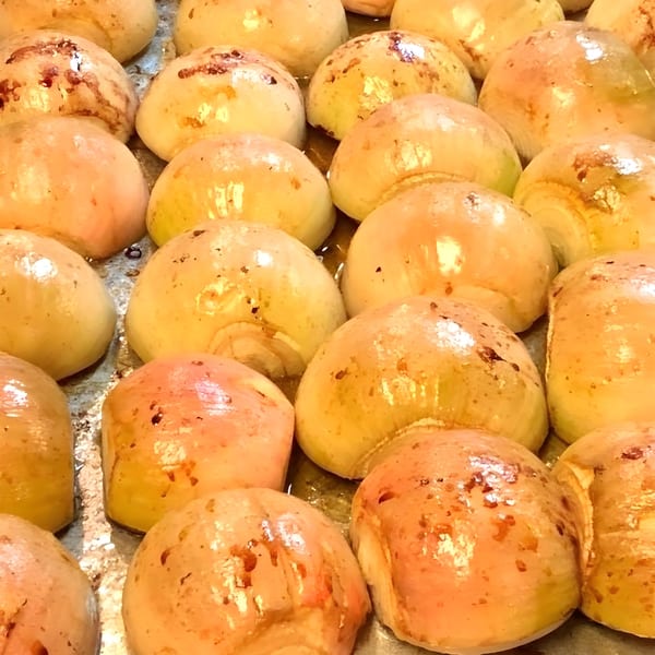 Close-up of many glistening, whole roasted onions with some browning, in a metal pan.